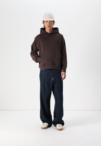 Brown sweatshirt with a hood, front pocket, loose-fit dark denim jeans, and white sneakers with light brown soles. Model wears a light cap.