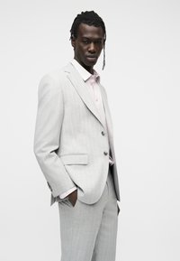 Homme aux cheveux tressés portant un costume gris clair à fines rayures et une chemise rose pâle, debout avec une main dans la poche, sur un fond blanc.