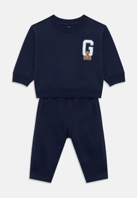 Marineblå baby tracksuit med lange ermer og bukser, med et hvitt bokstav "G"-merke og en liten teddy-bjørn på brystet.