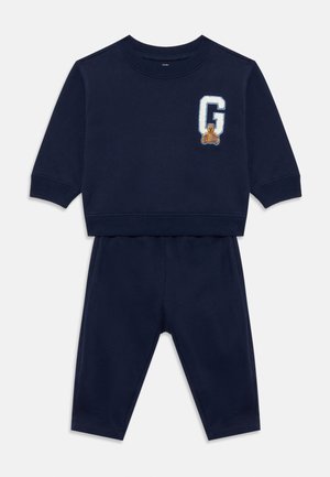 Survêtement pour bébé bleu marine avec des manches longues et un pantalon, arborant un patch de la lettre "G" blanche et un petit ours en peluche sur la poitrine.