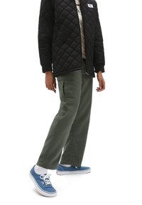 Chaqueta negra acolchada con pu ños de canalé, pantalones cargo verde oliva con bolsillos laterales y zapatillas de lona azules con cordones blancos.