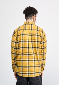 Camisa de franela amarilla con patrón de cuadros en negro y azul, de mangas largas, puños abotonados y dobladillo recto. Vista trasera que muestra el cuello y la costura del yugo.