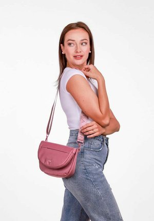 Růžová crossbody taška s designem zakřiveného klapky, nastavitelné ramenní pásmo s textem a hladkým povrchem. Nositá přes bílé tričko a džíny.