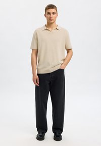 Polo en tricot beige à manches courtes et col, associé à un jean large noir et des chaussures noires, se tenant devant un fond neutre.