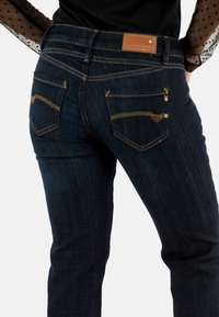 Jeans de mezclilla azul oscuro con costuras amarillas, que cuentan con dos bolsillos traseros y un parche de cuero marrón de la marca en la cintura.