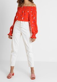Červená off-the-shoulder blůza s květinovým potiskem, nařaseným výstřihem a dlouhými rukávy, spárovaná s bílými džínami s vysokým pasem a růžovými sandály na blokovém podpatku.