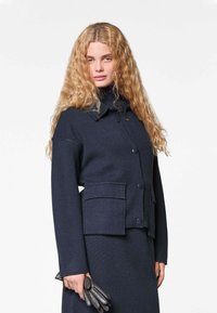 Navy wollen blend jas met een hoge hals, knoopsluiting, twee opgestikte zakken en geribde manchetten. Verfraaid met een trekkoorddetail.