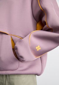 Lila sweatshirt med gula detaljer, som har en framficka och en logotyp på ärmen. Slät textur och avslappnad passform.