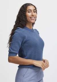 Blauer Strickpullover mit strukturierter Oberfläche, kurzen Ärmeln und gerafften Schulterdetails. Getragen mit hellgrauen Hosen.