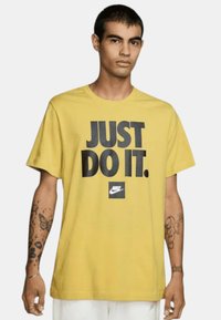 Camiseta amarilla de Nike con el emblemático texto negro "JUST DO IT." en negrita, logotipo cuadrado en el dobladillo, mangas cortas y un corte relajado.