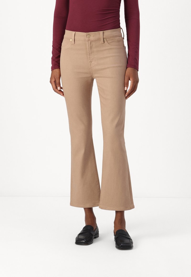 Personne portant un pantalon évasé beige, des mocassins noirs et un haut à manches longues bordeaux, debout devant un fond blanc uni.