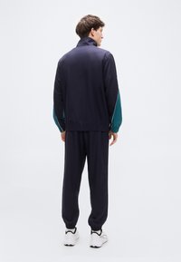 Ανδρική αθλητική φόρμα σε ναυτικό μπλε με τιρκουάζ λεπτομέρειες. Διαθέτει ψηλό γιακά, ελαστικά τελειώματα στα μανίκια και εφαρμοστά παντελόνια. Κατασκευασμένη από λείο ύφασμα.