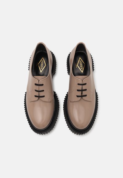 Beige leather shoes with three black lace eyelets, rubber textured sole, and black serrated accents. Logo on black insole visible.