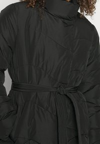 Cappotto puffer nero con alto colletto, vita con cintura e motivo trapuntato per una maggiore texture e calore. Tessuto esterno liscio e lucido.