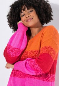 Donna con capelli ricci che indossa un maglione lavorato a maglia di un vivace arancione, rosso e rosa, sorridendo con gli occhi chiusi e la mano appoggiata sulla guancia.