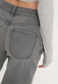 Vaqueros de tiro alto en gris con una textura suave, que presentan un bolsillo trasero y detalles de costura en contraste. Se llevan con un top de manga larga en gris claro.