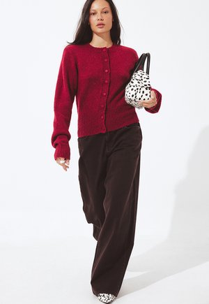 Rode gebreide cardigan met ronde hals en knoopsluiting aan de voorkant, gecombineerd met donkere wijde broek en een zwart-witte gespikkelde handtas.
