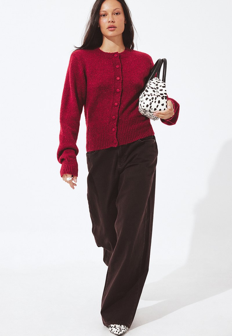 Rode gebreide cardigan met ronde hals en knoopsluiting aan de voorkant, gecombineerd met donkere wijde broek en een zwart-witte gespikkelde handtas.