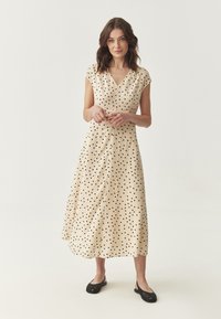 Beige midi-jurk met zwarte stippen, v-hals, korte mouwen en een vloeiende uitstraling. Gepaarde met zwarte platte schoenen, soepele stof.