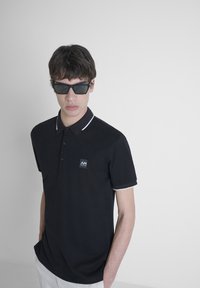 Crna polo majica s bijelim detaljima, s malim logom na lijevoj strani prsiju, izrađena od teksturirane tkanine, kratkih rukava i ovratnika.