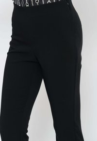 Primo piano di una persona che indossa pantaloni neri aderenti e una maglietta nera con un motivo a catena bianca, in piedi contro uno sfondo chiaro uniforme.