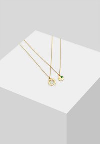 Goldene Halsketten mit zwei Anhängern: einem blumenförmigen Anhänger mit ausgeschnittenem Design und einem kleineren herzförmigen Anhänger mit grünem Akzent.