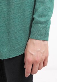 Teal neulepaita, jossa on raidalliset hihansuut, esitetty ranteen tasolla. Tasainen rakenne, löysä istuvuus ja hieman pidempi takasauma.