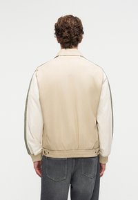 Mann mit lockigem Haar, der eine beige Jacke mit weißen und olivfarbenen Ärmeln sowie dunkelgrauen Jeans trägt und vor einem weißen Hintergrund steht.