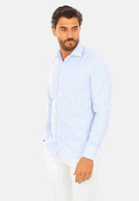 Camicia button-down a righe blu chiaro e bianco con maniche lunghe e colletto, realizzata in tessuto liscio. Il modello si trova in posizione casual con le mani nelle tasche.