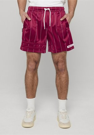Mann trägt burgunderrote Satin-Pinstripe-Shorts mit weißem Kordelzug, weiße Sportsocken und weiße Turnschuhe, steht mit den Händen in den Taschen.