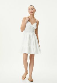 Abito bianco con corsetto aderente, vita smock e gonna a strati svasata. Presenta spalline sottili e tessuto testurizzato. Indossato con sandali dorati.