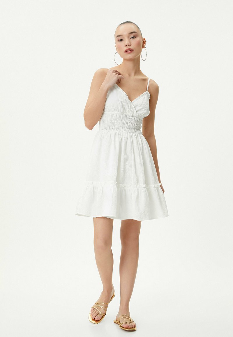 Abito bianco con corsetto aderente, vita smock e gonna a strati svasata. Presenta spalline sottili e tessuto testurizzato. Indossato con sandali dorati.