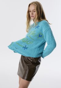 Türkiser, flauschiger Pullover mit grünen und blauen grafischen Mustern, kombiniert mit einem braunen Leder-Minirock. Gemütliche Textur und lockere Passform.