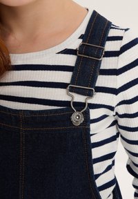 Salopette in denim con clip metallici regolabili, caratterizzata da cuciture arancioni e una maglietta aderente a righe in bianco con righe blu navy.