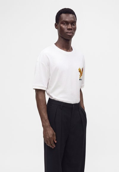 Junger Mann, der ein weißes T-Shirt mit einem kleinen gelben Adler-Design auf der Brust und schwarze plissierte Hosen trägt, steht vor einem weißen Hintergrund.