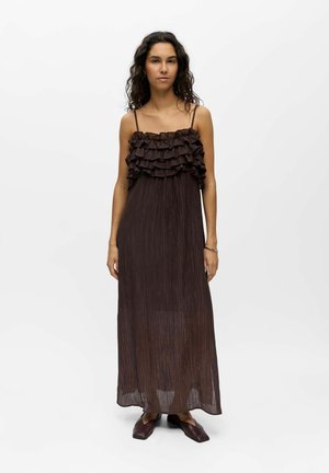 Femme debout portant une robe longue marron foncé sans manches avec un corsage à volants et des chaussures marron à bout carré sur un fond blanc uni.