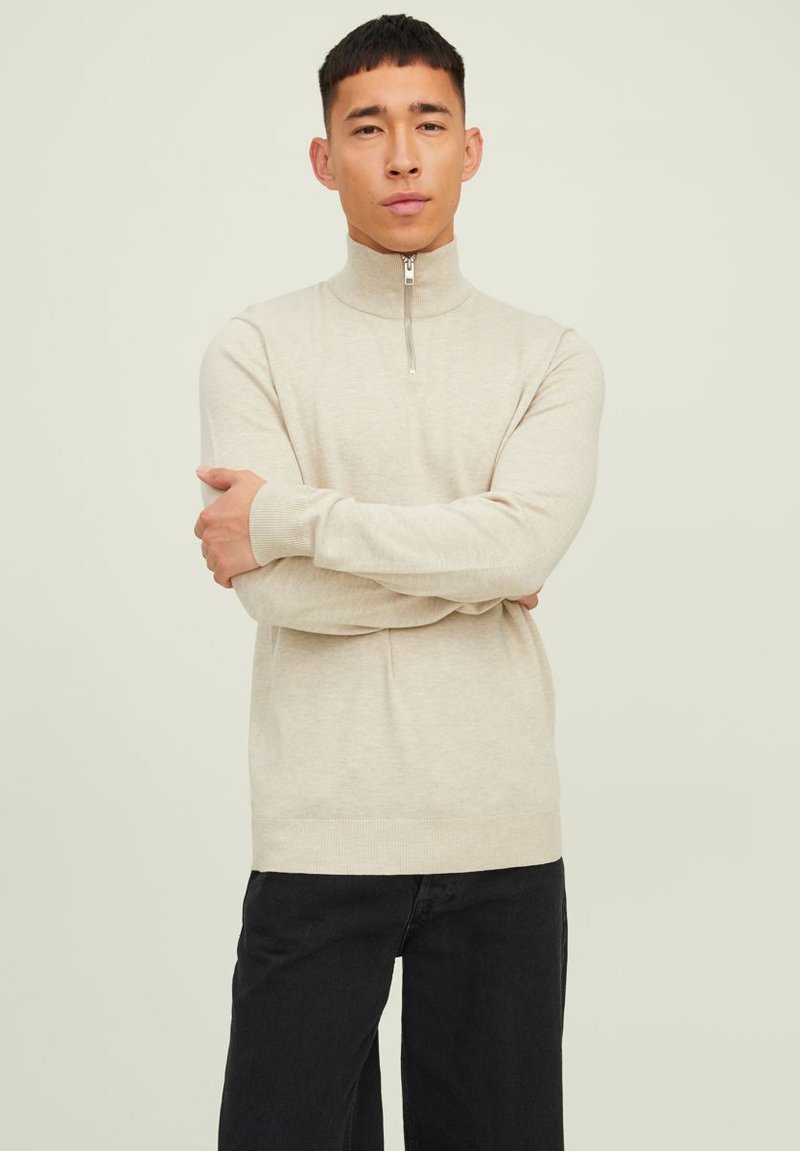 Jersey de cuello alto color crema con cuello con cremallera, puños y dobladillo acanalados. Lleva pantalones oscuros; textura suave, diseño ajustado.