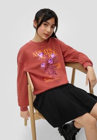 Un sweat-shirt rouge avec un motif floral et du texte, associé à une jupe plissée noire et des bottes de combat noires, assis sur une chaise en bois.