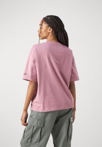 T-maica kratkih rukavov z okroglim izrezom v mehkem rožnatem bombažu, ki ima minimalno blagovno znamko na levi rokavi; v kompletu z ohlapnimi zelenimi vojaškimi hlačami.