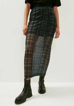 Femme portant une longue jupe transparente à carreaux avec des bottines noires épaisses et un haut noir, debout devant un fond clair uni.