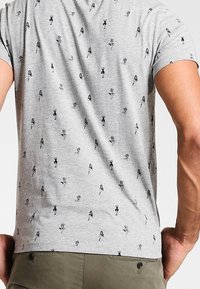 Šedé tričko s krátkým rukávem s opakujícím se černým grafickým vzorem malých postav. Texturovaná bavlněná tkanina s designem crew neck.