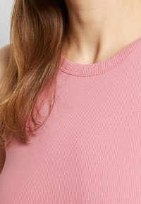 Gros plan d'une personne portant un haut sans manches rose côtelé, avec de longs cheveux bruns partiellement visibles sur l'épaule.