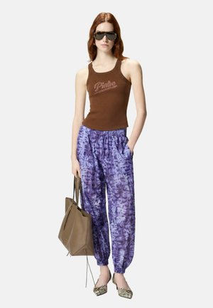 Donna con top marrone senza maniche, pantaloni jogger viola con fantasia, tacchi in pelle di serpente, occhiali da sole oversize, che tiene una borsa beige.