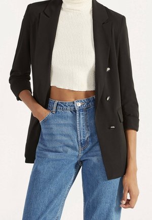Kvinde iført en sort blazer over en hvid ribbet cropped rullekrave og blå højhustede jeans med den ene hånd i lommen.