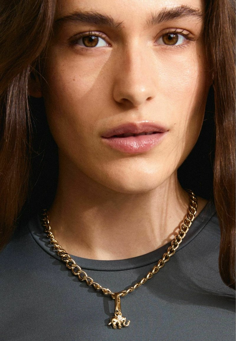 Collana in oro con design a maglie spesse e un ciondolo a forma di elefante; indossata sopra un top scuro e texturizzato.