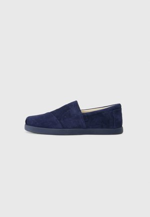 Sapatos slip-on azul-marinho com uma textura de camurça suave, apresentando um design assimétrico e um bico arredondado. Confeccionados com uma sola de borracha flexível.