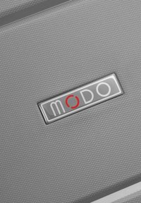 Superficie di una valigia grigia texsturata con una prominente placca di logo argentato con il nome del marchio "modo" e un accento rosso nella lettera "o".