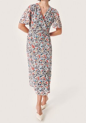 Vrouw die een kleurrijke bloemen midi wikkeljurk met korte mouwen en witte puntige hakken draagt, staand op een lichte achtergrond.