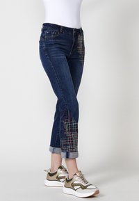 Denim kavbojke temnega pranja z barvitimi vezenimi detajli na levem stegnu in zavihanimi manšetami. Kombinirane s športnimi supergami.