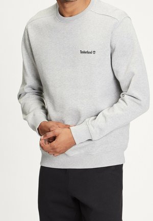 Mand iført lysegrå Timberland sweatshirt med lange ærmer og sorte bukser, hænderne foldet foran.